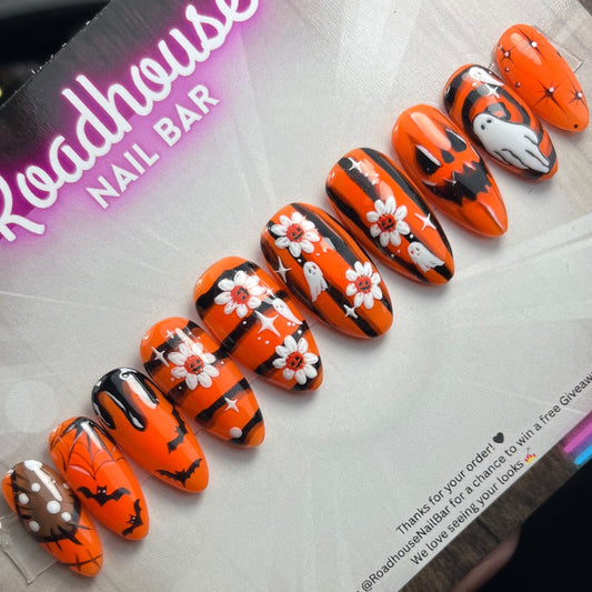 Hand-painted Halloween press-on nails with orange and black spooky designs, featuring ghosts, bats, pumpkins, spider webs, and flowers.