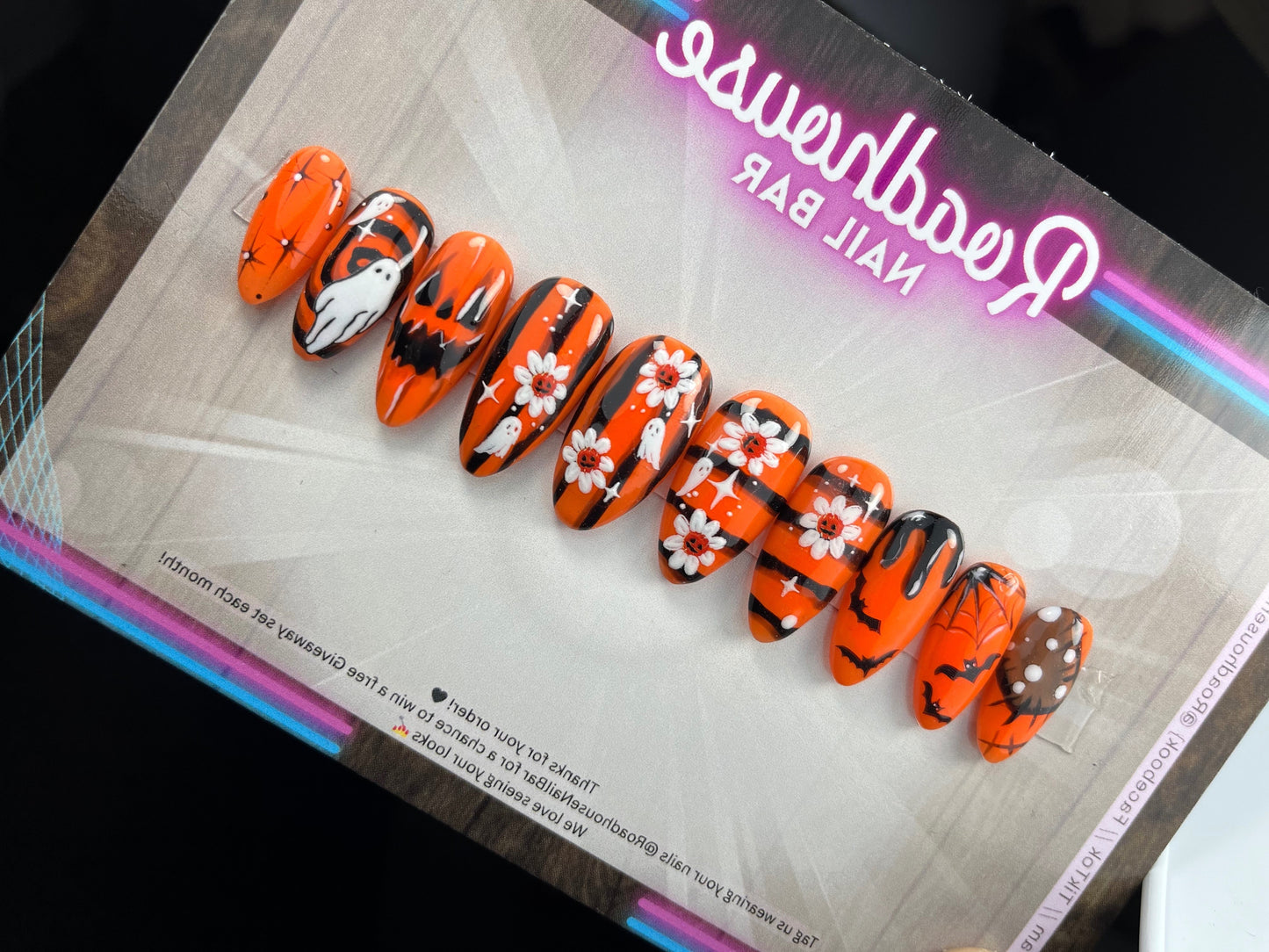 Hand-painted Halloween press-on nails with orange and black spooky designs, featuring ghosts, bats, pumpkins, spider webs, and flowers.