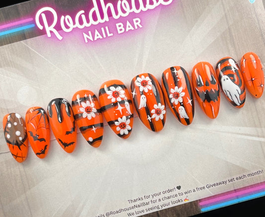 Hand-painted Halloween press-on nails with orange and black spooky designs, featuring ghosts, bats, pumpkins, spider webs, and flowers.