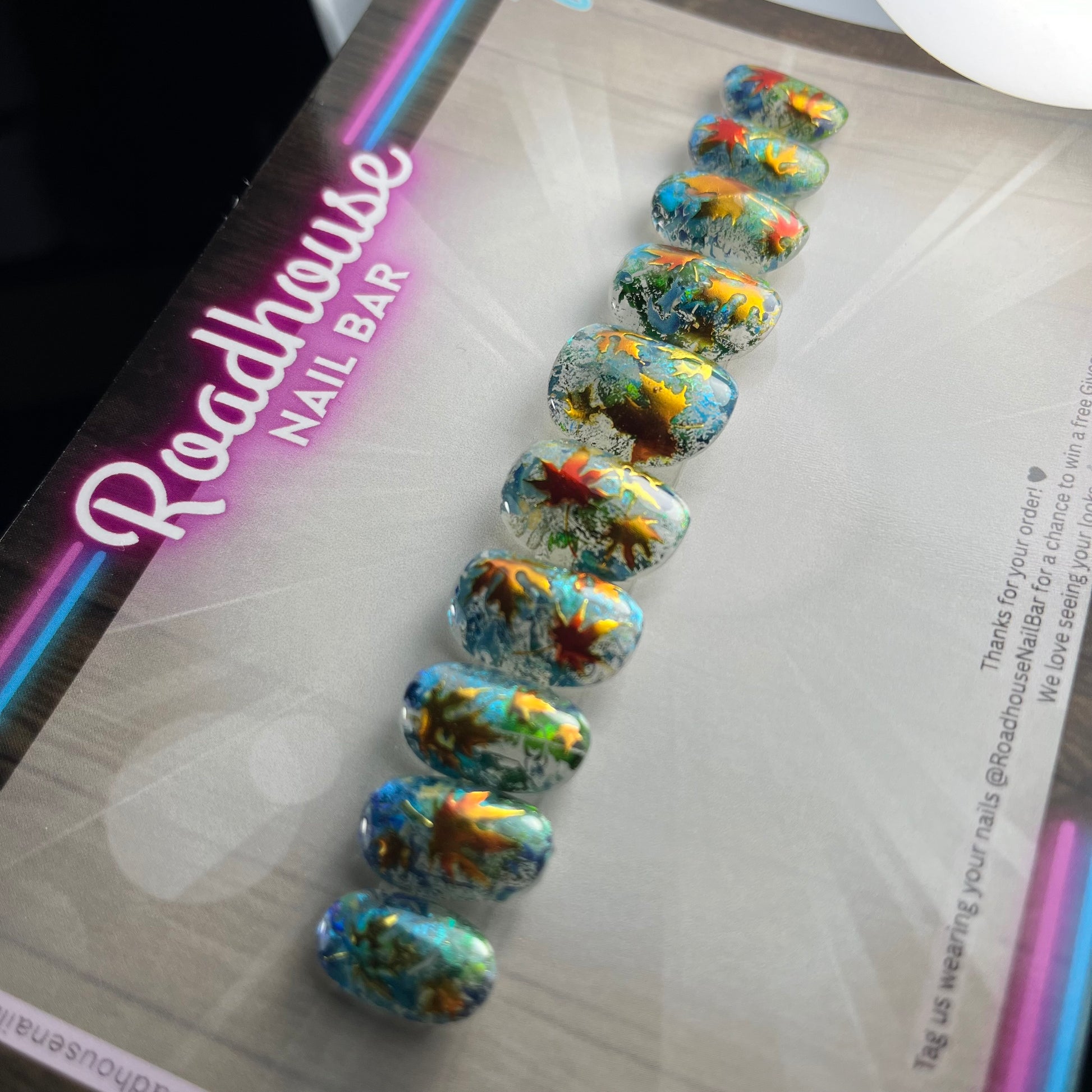 Hand-painted press-on nails with glass-effect base, aqua foiling, and metallic copper autumn leaves, displayed on Roadhouse Nail Bar backing card.