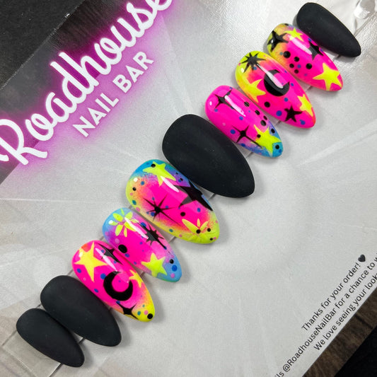 A set of false press-on nails in a retail package from Roadhouse Nail Bar. The set includes five black almond-shaped nails and five neon-patterned stiletto nails with pink, yellow, blue, and green colors, black stars, and crescent moon designs.