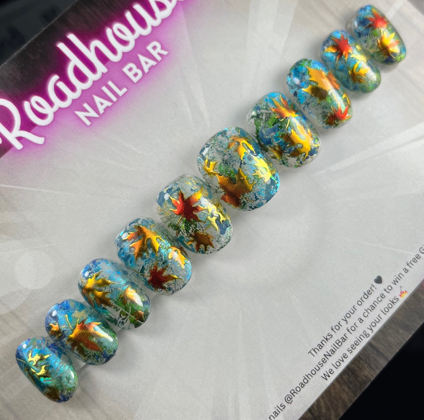 Hand-painted press-on nails with glass-effect base, aqua foiling, and metallic copper autumn leaves, displayed on Roadhouse Nail Bar backing card.