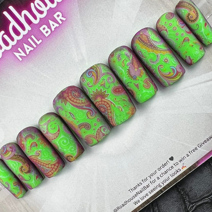 A set of press-on nails with a green and brown paisley pattern, displayed on their retail card which is labeled 'Roadhouse Nail Bar'. The card is placed on a dark textured surface.