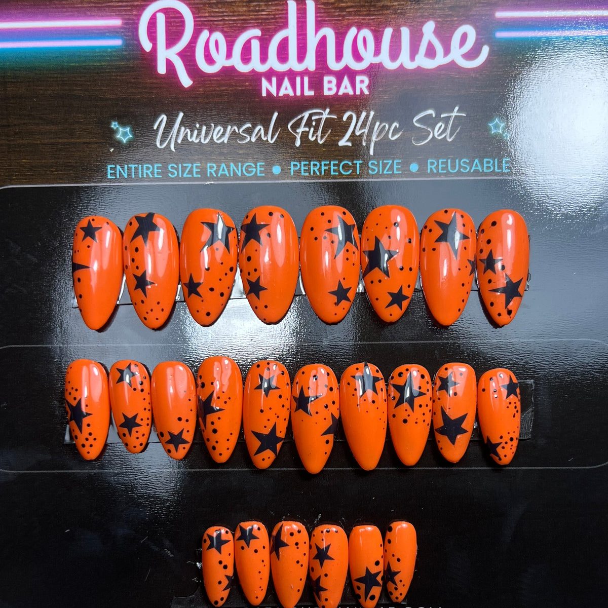 A 12-piece set of orange press-on nails with black star patterns, presented in a black retail box labeled 'Roadhouse Nail Bar'.
