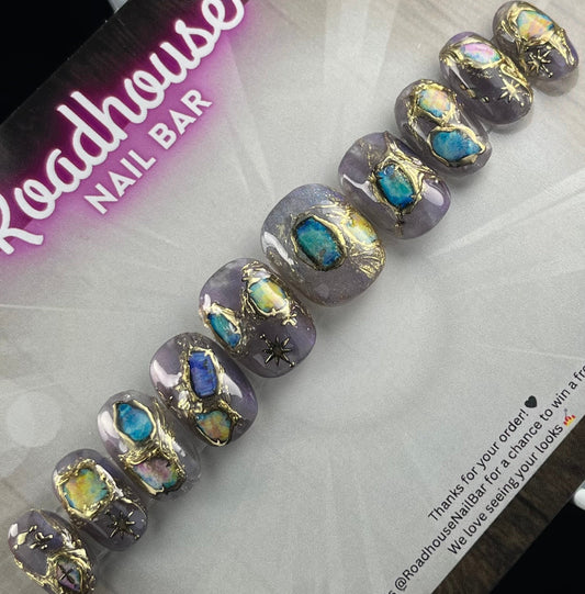 Short natural-oval press-on nails in lavender-grey with hand-painted opal-effect gemstones, molten gold detailing, and celestial starburst accents, displayed on Roadhouse Nail Bar backing card.