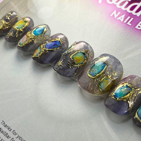 Short natural-oval press-on nails in lavender-grey with hand-painted opal-effect gemstones, molten gold detailing, and celestial starburst accents, displayed on Roadhouse Nail Bar backing card.