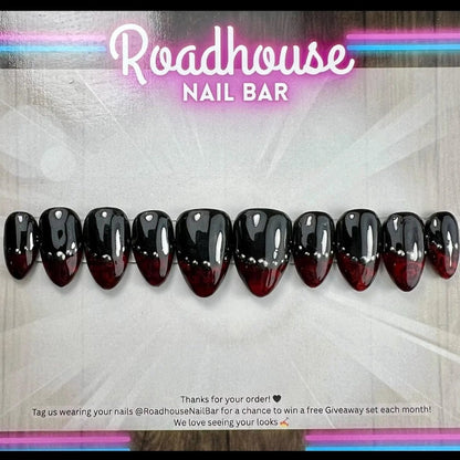 A set of 12 glossy press-on nails displayed horizontally, featuring a black-to-deep-red ombre gradient with silver chrome accents. The nails are presented on a branded background card for 'Roadhouse Nail Bar' with promotional text at the bottom.