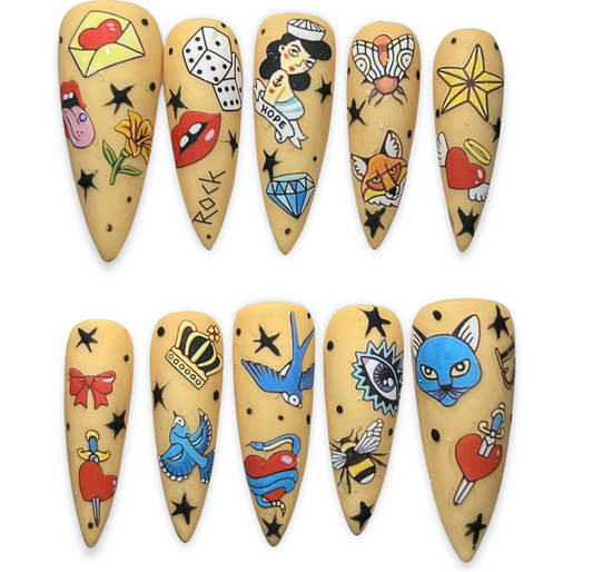 Traditional tattoo-inspired press-on nails with old-school tattoo art on a neutral base, including swallows, hearts, skulls, and pin-up motifs.